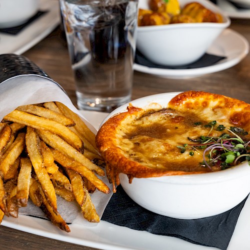A plate of crispy fries next to a hot bowl of French onion soup, topped with cheese and herbs, on a table with drinks in the background.