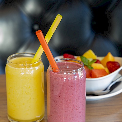 Two colorful smoothies in jars with straws (yellow and pink) sit on a wooden counter, fruit bowl in background.
