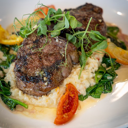 A plated steak atop creamy risotto with greens and cherry tomatoes, garnished with microgreens — a savory, restaurant-style presentation.
