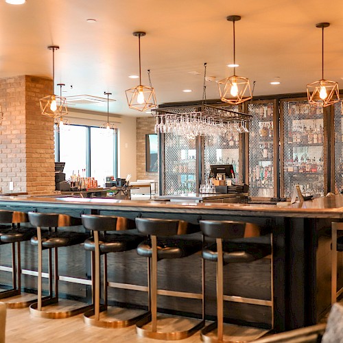 A stylish bar area with a long counter, high stools, hanging pendant lights, brick accents, and shelves of glassware in a warm, inviting setting.