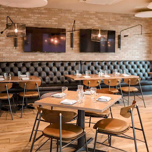 A modern restaurant interior with wooden tables and tan chairs, a long tufted black leather banquette, wall sconce lighting, and place settings ready for guests.