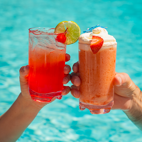 Two hands hold up tropical cocktails by a bright pool, red and peach drinks with fruit garnishes, to celebrate sunny vibes.