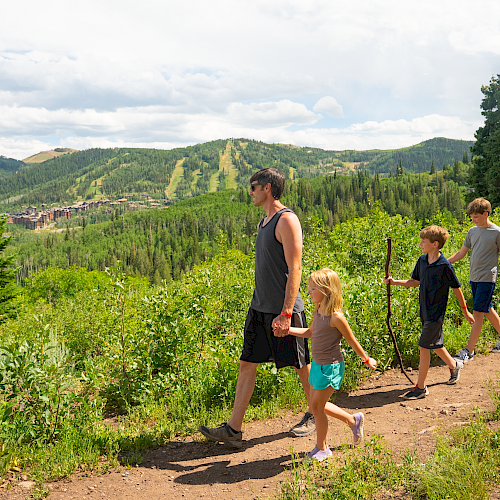 Family hike along a dirt trail in a lush mountainside landscape, kids exploring with adults under a partly cloudy sky.