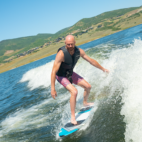 A man wearing a life vest waterskis on a lake, riding a wave with hills in the background, sunny day, blue sky, action pose.