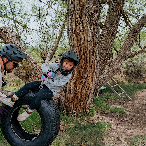 Two people share a playful moment outdoors: a child on a tire swing tied to a sturdy tree while an adult helps push, smiles all around.