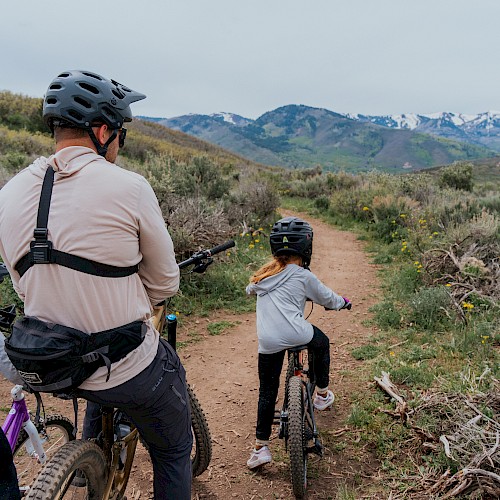 Three people bike along a dirt trail in a mountain landscape, wearing helmets and casual gear, as distant snow-capped hills rise ahead.