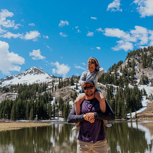 A couple enjoys a mountain lake scene: a man carries a woman on his shoulders, with snow-capped peaks, evergreen trees, and a bright blue sky behind them.