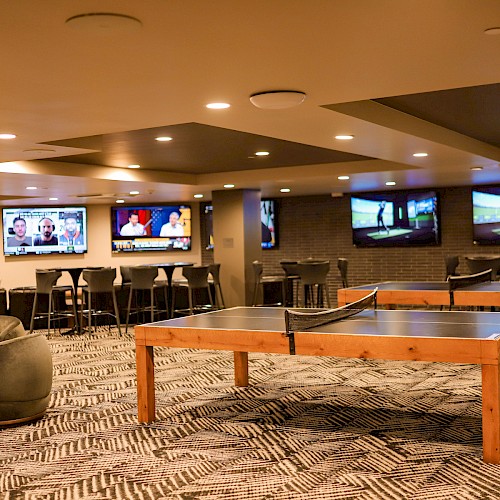 A spacious game lounge with ping-pong tables, seating areas, and multiple TVs on the walls for viewing events.