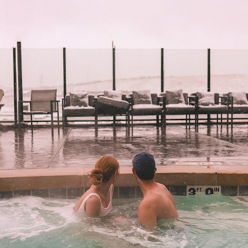 Two people sit close in a pool, facing away from the camera, chatting by the water while lounge chairs and a glass fence sit beyond. ending with a period.