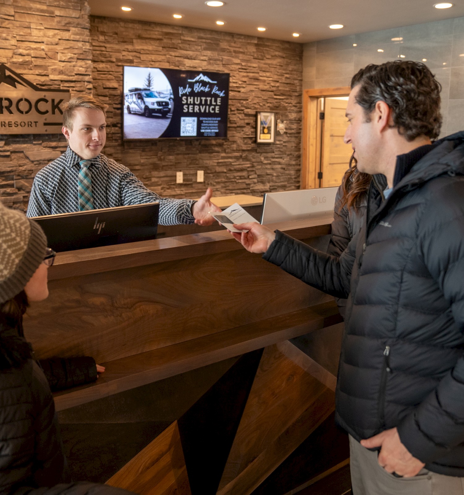 Three people at a Black Rock open-lobby counter; receptionist behind desk hands someone a paper, snowy jackets indoors.