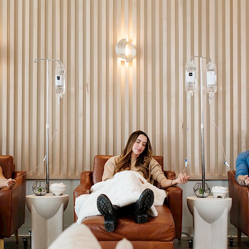 Three people sit in a modern lounge with IV stands behind them, each looking relaxed and patient, blankets over laps, casual vibe.