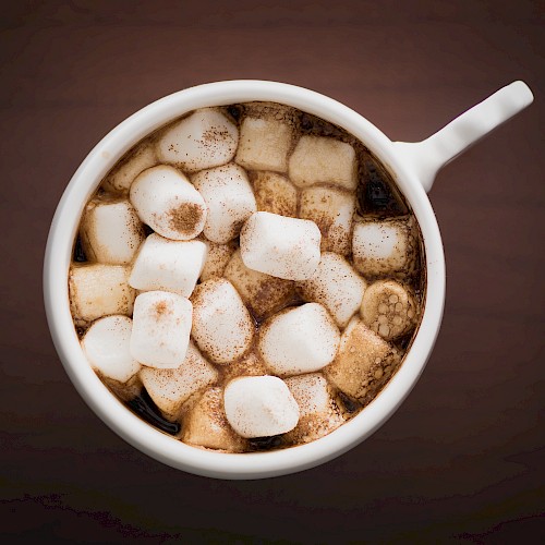 A white mug filled with hot chocolate topped with fluffy marshmallows, snowy squares dusted with cocoa.