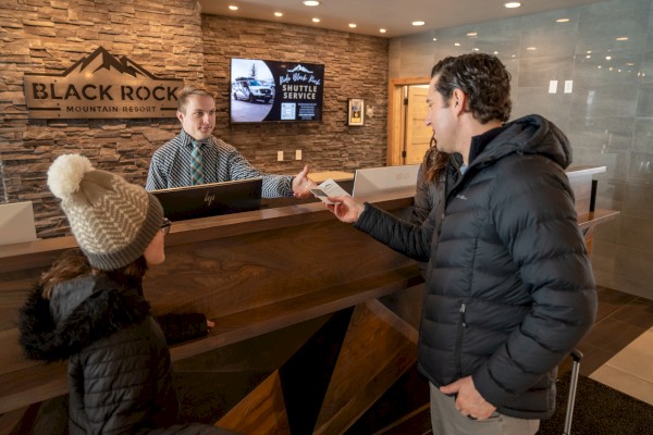 Two people check in at a hotel reception desk with a receptionist handing a key; brick wall, “Black Rock” sign, and screens behind.