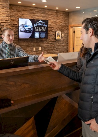 A man at a rustic front desk greets two visitors in a ski-theme lobby, with a “Black Rock Mountain Resort” sign and stone walls.