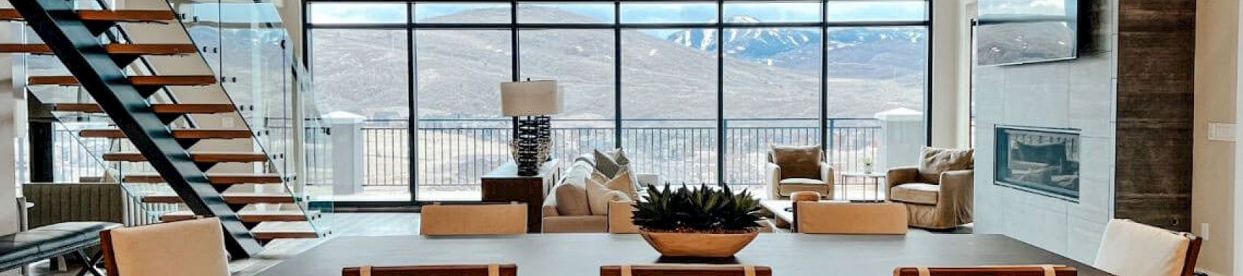 A modern living room with large windows, mountain views, a staircase, dining table, chairs, sofas, and a TV on the wall, ending the sentence.