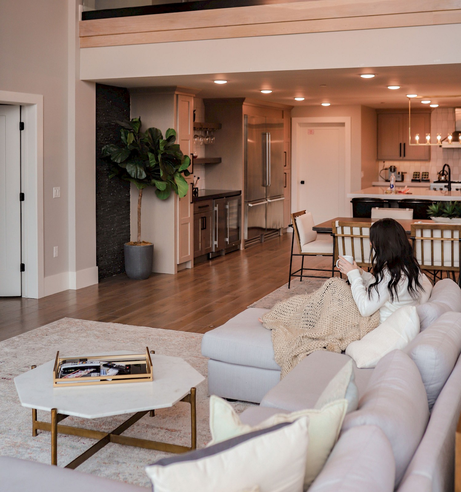 A modern, open-concept living area with a high ceiling, balcony, and stylish gray sectional sofa; cozy seating and a kitchen island in the background.
