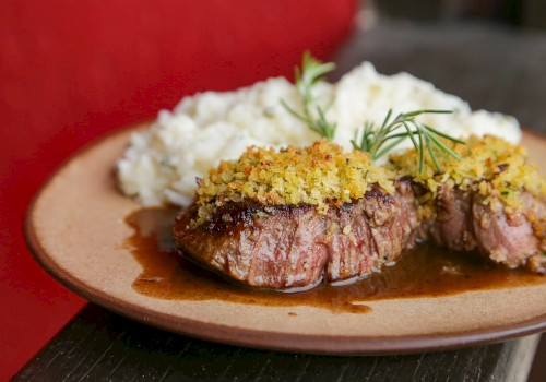 A plate with two slices of medium-rare steak topped with crispy breadcrumbs and a lemony herb sauce, garnished with dill.