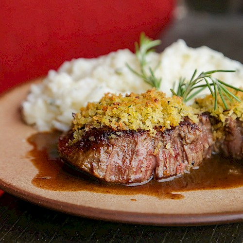 A plate with two slices of medium-rare steak topped with crispy breadcrumbs and a lemony herb sauce, garnished with dill.