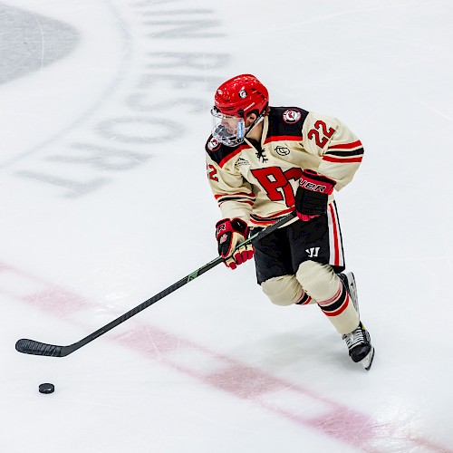 A hockey player in jersey number 22, wearing a red helmet, controls the puck on the ice rink.