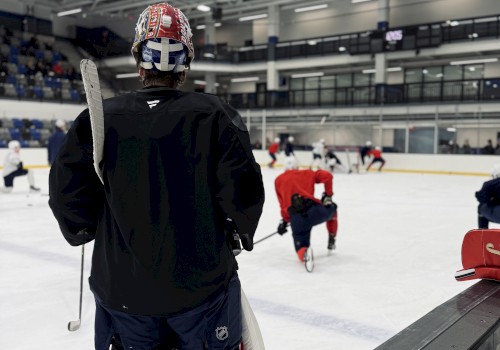 A hockey goalie in black gear stands on the rink watching players practice on an indoor ice hockey rink.