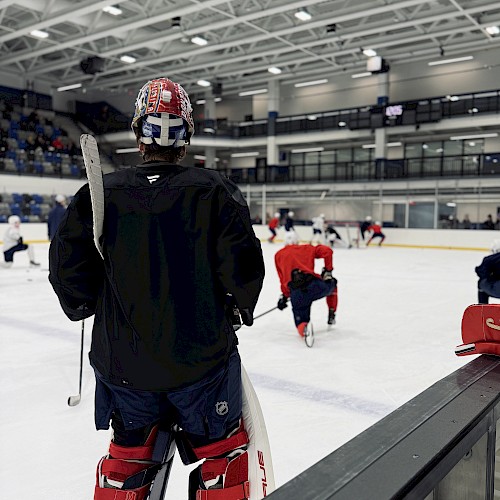 A hockey goalie in black gear stands on the rink watching players practice on an indoor ice hockey rink.